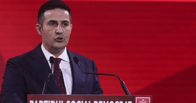 Un bărbat cu părul scurt, purtând costum închis la culoare, cămașă albă și cravată închisă, vorbește la un podium cu microfoane în fața unui fundal roșu.