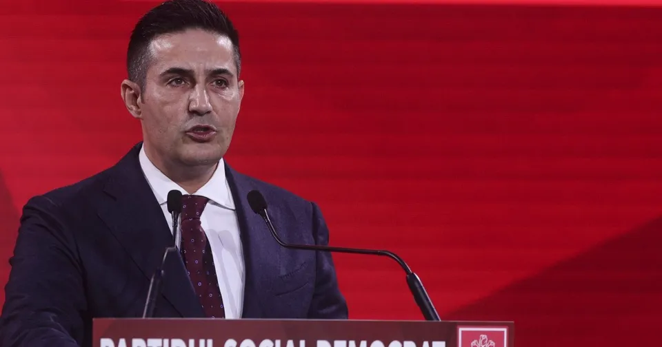 Un bărbat cu părul scurt, purtând costum închis la culoare, cămașă albă și cravată închisă, vorbește la un podium cu microfoane în fața unui fundal roșu.