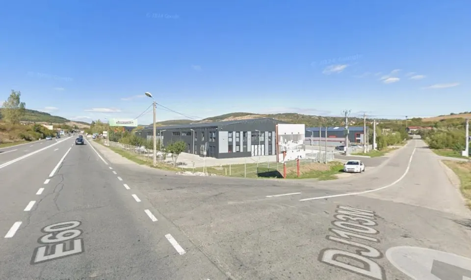 Imagine panoramică a unei intersecții rutiere, cu un drum principal marcat E60 și un drum secundar marcat DJ103M, sub un cer senin, cu clădiri și dealuri în fundal.