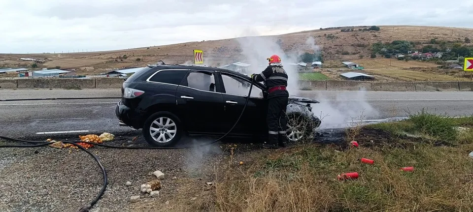 Un pompier în uniformă neagră cu detalii roșii stă lângă un autoturism negru de care se ridică fum pe marginea unui drum asfaltat.