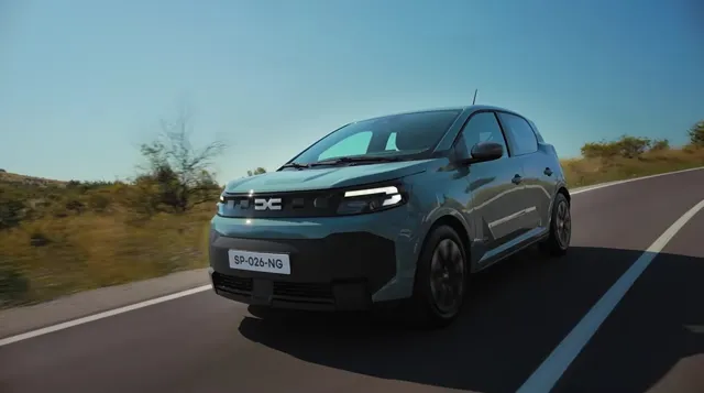 Un autovehicul compact de culoare gri închis circulă pe un drum asfaltat sub un cer senin.