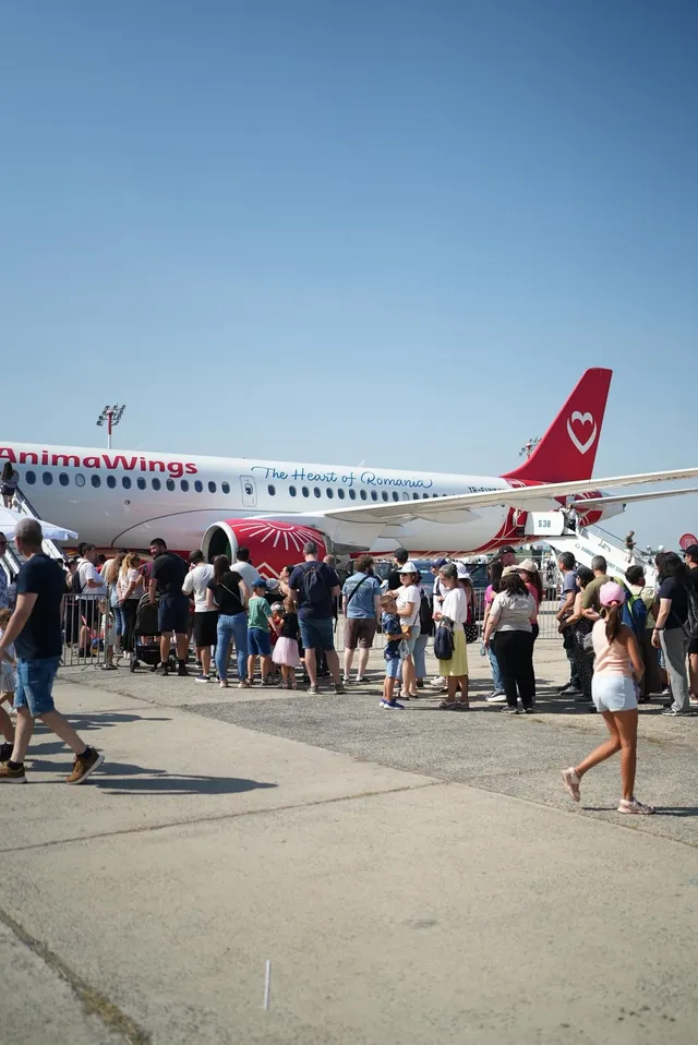 O mulțime de oameni stă la coadă pe o pistă de beton lângă un avion alb cu marcaje roșii sub un cer senin.