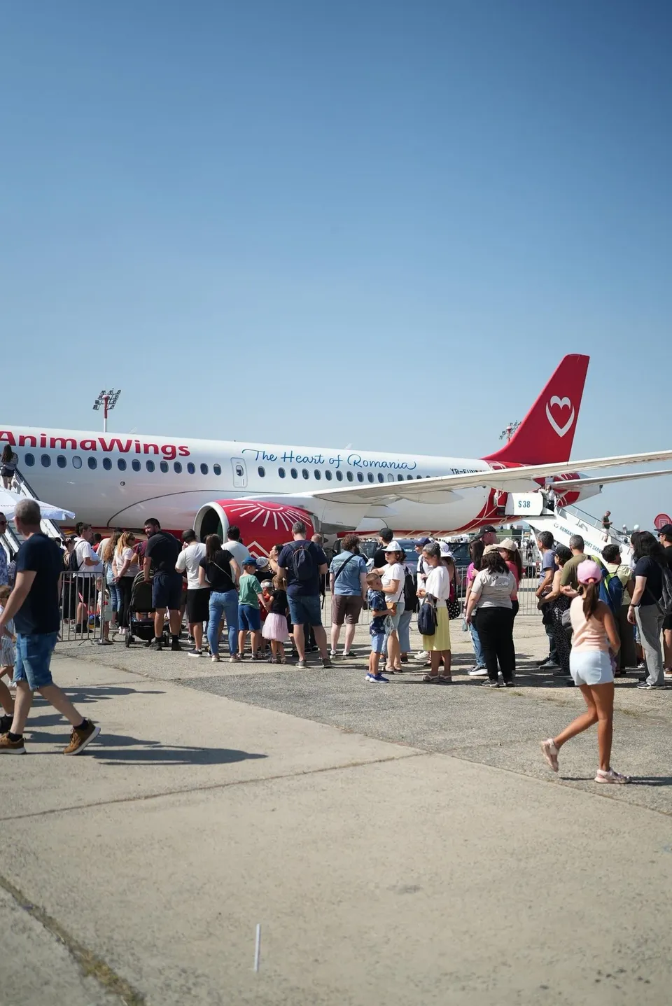 O mulțime de oameni stă la coadă pe o pistă de beton lângă un avion alb cu marcaje roșii sub un cer senin.