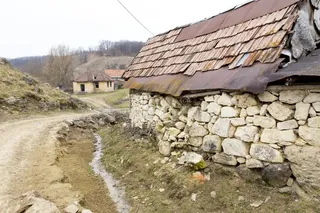 O casă cu pereți din piatră și acoperiș de țiglă uzate se află lângă un drum de țară într-un peisaj rural.