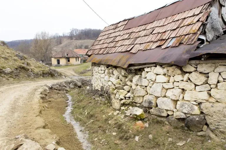 O casă cu pereți din piatră și acoperiș de țiglă uzate se află lângă un drum de țară într-un peisaj rural.