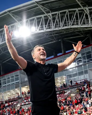Un bărbat cu mâinile ridicate poartă un tricou negru și se află într-un stadion cu tribune ocupate de oameni în fundal.