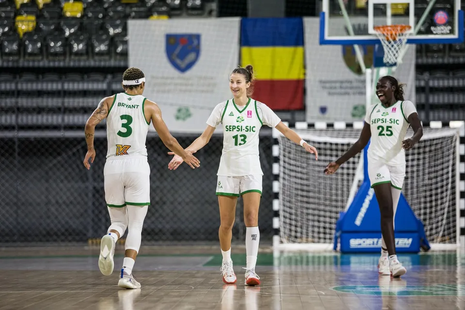 Trei jucătoare de baschet cu echipamente albe și verzi stau pe un teren de sport în fața unui coș de baschet și a unei ținte de fotbal.
