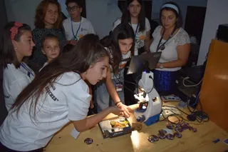 Un grup de copii și adolescenți privește atent o mostră aflată pe o masă lângă un microscop într-o încăpere cu lumină slabă.