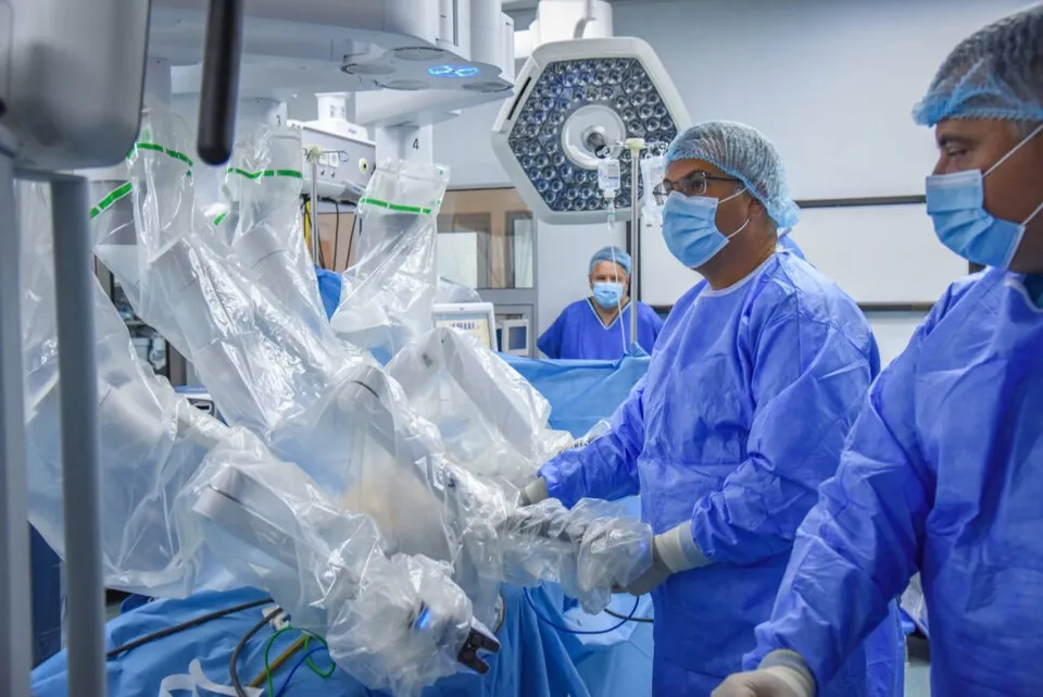 Trei medici în halate și măști de protecție albastre operează lângă un aparat robotic acoperit cu folie transparentă într-o sală de operație.