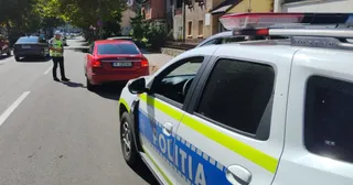 Un polițist cu vestă reflectorizantă stă pe marginea drumului lângă un autoturism roșu, în timp ce o mașină de poliție albă este parcată în prim-plan.