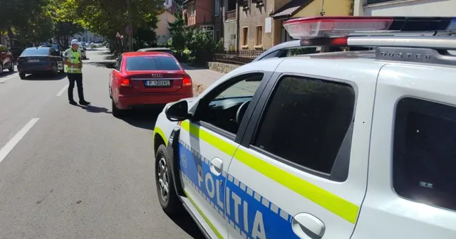 Un polițist cu vestă reflectorizantă stă pe marginea drumului lângă un autoturism roșu, în timp ce o mașină de poliție albă este parcată în prim-plan.