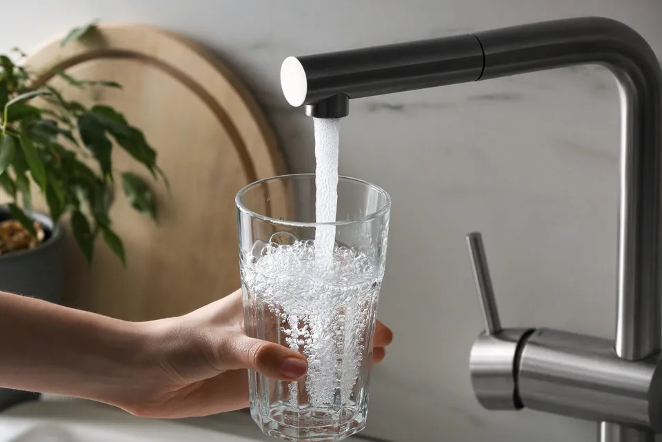 O mână ține un pahar transparent sub un robinet metalic din care curge un jet de apă.