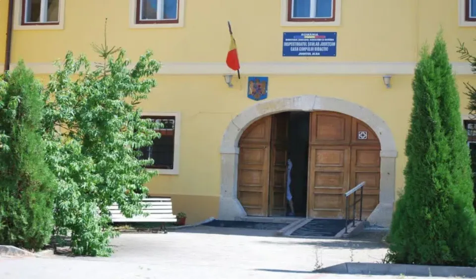 O clădire galbenă cu ferestre albe, un arc de intrare din piatră cu uși de lemn și vegetație de tip conifer în prim-plan.