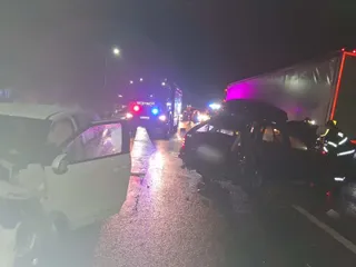 Două autoturisme și un camion sunt situate pe un drum umed noaptea, sub lumini de alertă colorate.