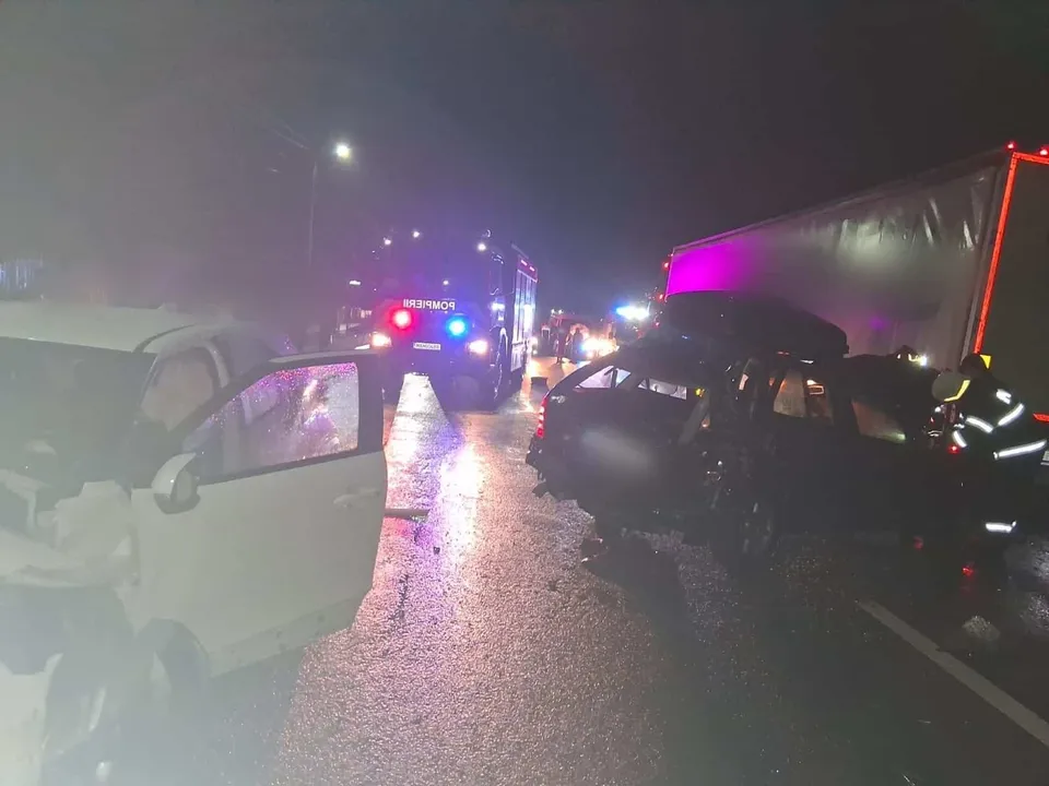 Două autoturisme și un camion sunt situate pe un drum umed noaptea, sub lumini de alertă colorate.