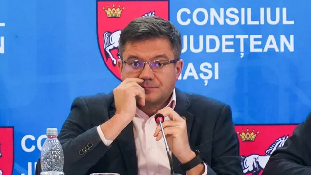 Un bărbat cu ochelari și sacou negru stă la o masă în fața unui panou albastru cu inscripții și un scut colorat.