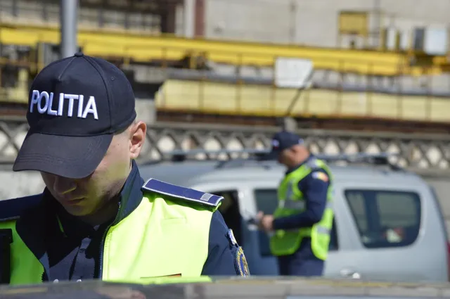 Un polițist cu șapcă neagră și vestă reflectorizantă verde stă în prim-plan, în timp ce un alt agent se află lângă un autoturism gri în fundal.