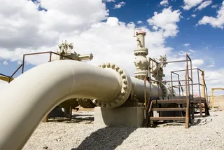 O conductă metalică mare, curbată, este conectată la un sistem de valve și supape într-o zonă deschisă sub un cer cu nori.