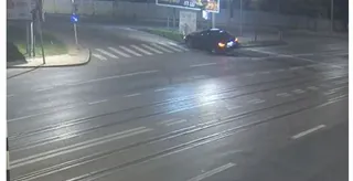 Un autoturism negru este poziționat pe marginea unei străzi asfaltate, lângă un trotuar și un stâlp, sub lumină nocturnă.