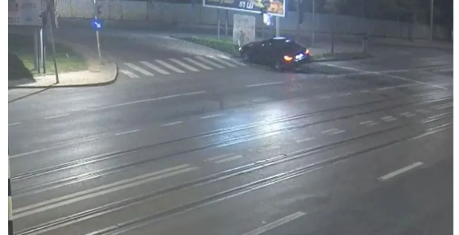 Un autoturism negru este poziționat pe marginea unei străzi asfaltate, lângă un trotuar și un stâlp, sub lumină nocturnă.