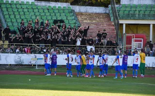 Un grup de jucători în echipamente albastre și albe stau pe teren, în timp ce suporteri în haine negre stau pe tribunele verzi ale unui stadion.