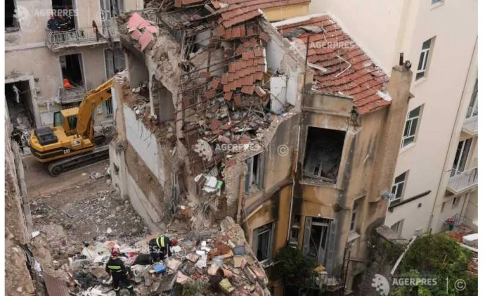 O excavatoare galbenă se află lângă o clădire parțial dărâmată cu resturi de cărămizi și beton împrăștiate pe sol.