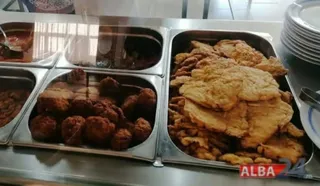 Mai multe tăvi metalice conțin diverse tipuri de alimente prăjite și gătite, dispuse într-o zonă de servire.
