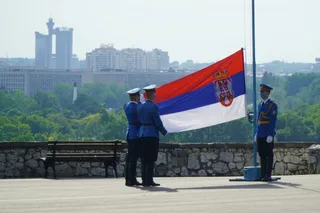 Trei bărbați în uniforme albastre stau pe o terasă lângă un stâlp cu un steag sârb, având clădiri și copaci în fundal.