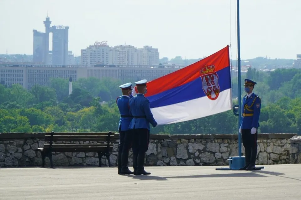 Trei bărbați în uniforme albastre stau pe o terasă lângă un stâlp cu un steag sârb, având clădiri și copaci în fundal.
