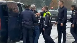 Mai mulți polițiști în uniformă întunecată și un bărbat în blugi și tricou negru stau lângă o mașină albastră deschisă.