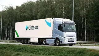 Un camion alb cu inscripția Girteka circulă pe o autostradă lângă o pădure de copaci verzi.