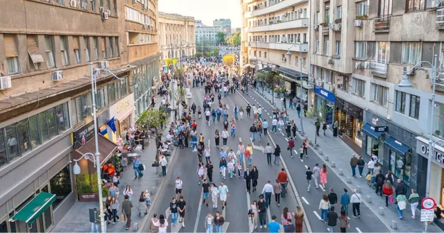 O mulțime de oameni merge pe un bulevard urban flancat de clădiri înalte și magazine pe ambele părți ale străzii.