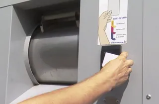 O mână ține un card alb lângă un cititor de carduri montat pe o suprafață gri metalică lângă o deschidere circulară.