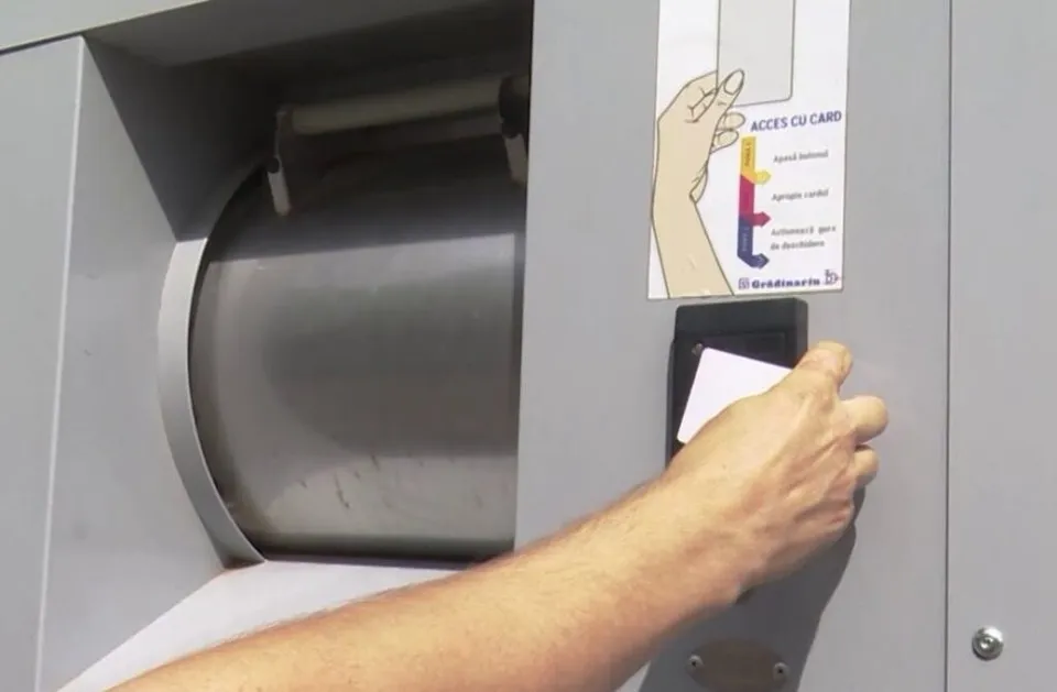 O mână ține un card alb lângă un cititor de carduri montat pe o suprafață gri metalică lângă o deschidere circulară.