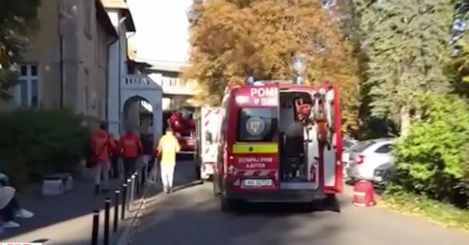 Un vehicul de intervenție roșu este parcat pe un drum, lângă persoane purtând haine de protecție și un vehicul alb.