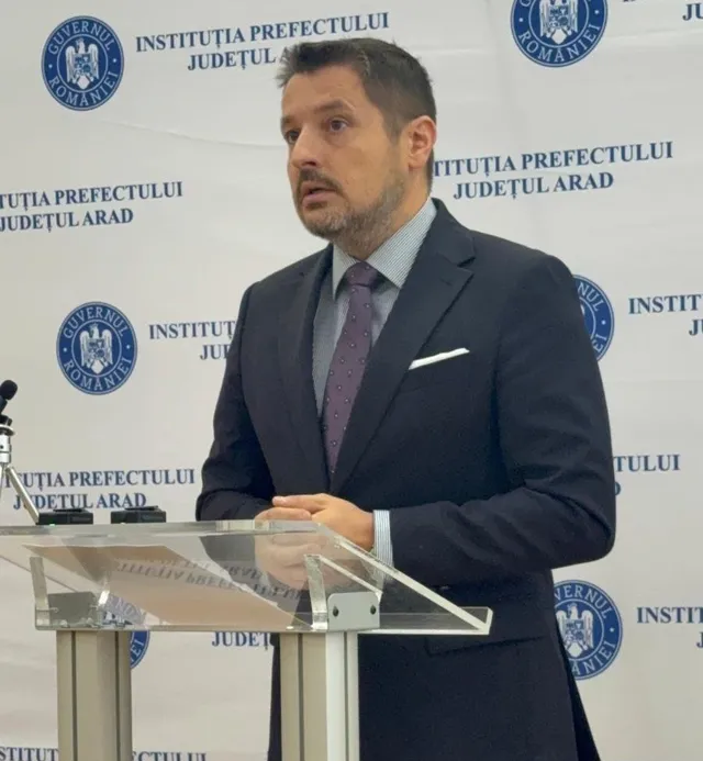 Un bărbat cu barbă, îmbrăcat în costum închis la culoare, cămașă gri și cravată mov, stă în spatele unui podium transparent în fața unui fundal alb cu logouri.