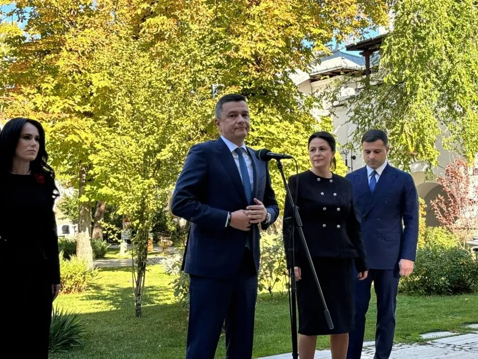Patru persoane în haine oficiale stau pe un covor verde în fața unor copaci cu frunze galbene și verzi lângă un microfon.