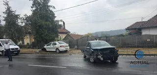 Un vehicul alb de poliție și o mașină neagră cu partea frontală distrusă sunt oprite pe un drum umed lângă un gard și copaci.