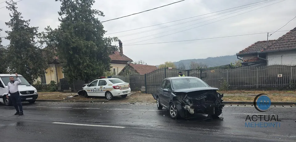 Un vehicul alb de poliție și o mașină neagră cu partea frontală distrusă sunt oprite pe un drum umed lângă un gard și copaci.