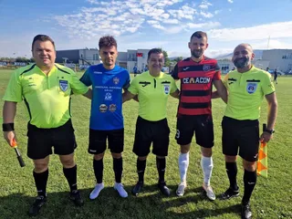 Cinci bărbați stau în linie pe un teren de fotbal sub un cer înnorat, purtând echipamente sportive și tricouri de arbitru de culoare galbenă.
