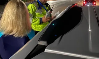 Un agent de poliție în uniformă reflectorizantă verde deschide o portieră de autoturism lângă o persoană cu părul blond într-un cadru nocturn.