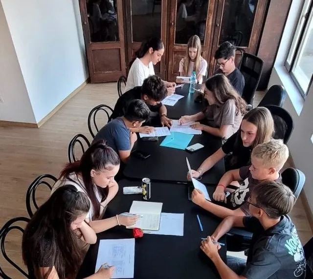 Un grup de copii și adolescenți stau la mese negre, scriind sau citind pe foi de hârtie într-o încăpere cu uși din lemn.