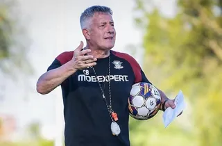 Un bărbat cu părul scurt și grij, purtând un tricou negru cu accente roșii, ține o minge de fotbal și o foaie de hârtie.