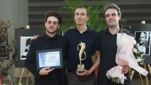 Trei bărbați stau în picioare, unul purtând un trofeu auriu, unul o placă de premiu și unul un buchet de flori.