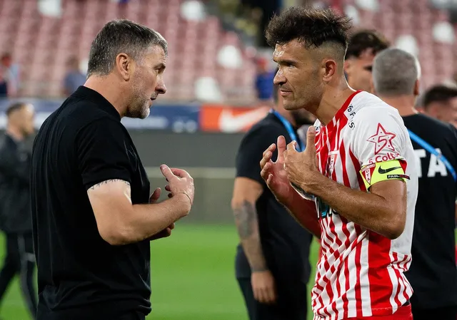 Un bărbat într-un tricou negru vorbește cu un jucător de fotbal îmbrăcat într-un echipament alb cu dungi roșii pe un teren de fotbal.
