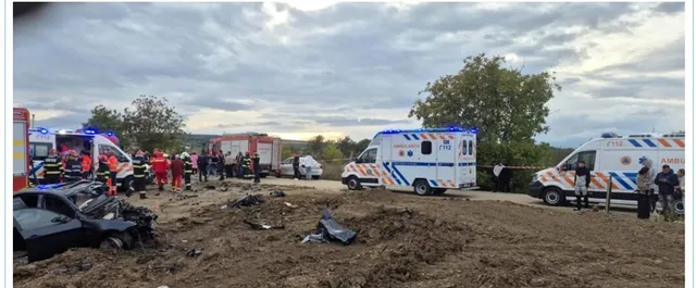 Vehicule de intervenție, inclusiv ambulanțe și o autospecială de pompieri, sunt parcate pe un teren de lângă un autoturism distrus într-un accident rutier.