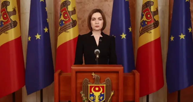 O femeie cu părul scurt și brunet stă în spatele unui podium de lemn în fața mai multor steaguri.