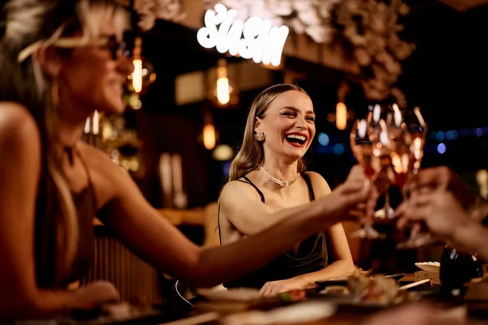 Două femei zâmbesc la o masă de restaurant, ridicând pahare cu vin într-un cadru cu lumini calde și un neon scris SUSHI.