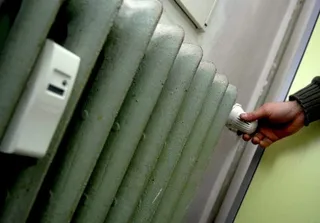O mână ține un termometul digital lângă un calorifer metalic de culoare verde deschis.