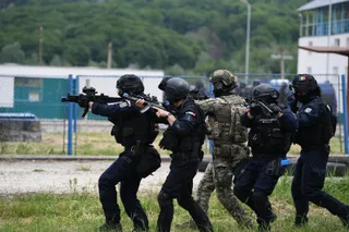 Cinci persoane în uniforme tactice și căști, purtând arme, se deplasează pe un teren cu iarbă în fața unor clădiri și copaci.